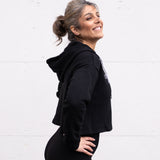 Woman standing facing sideways wearing a cropped bamboo-organic cotton fleece hoodie in black, featuring a finished hem, white drawstrings at the hood and the A$$piration Project logo on the front in light grey.