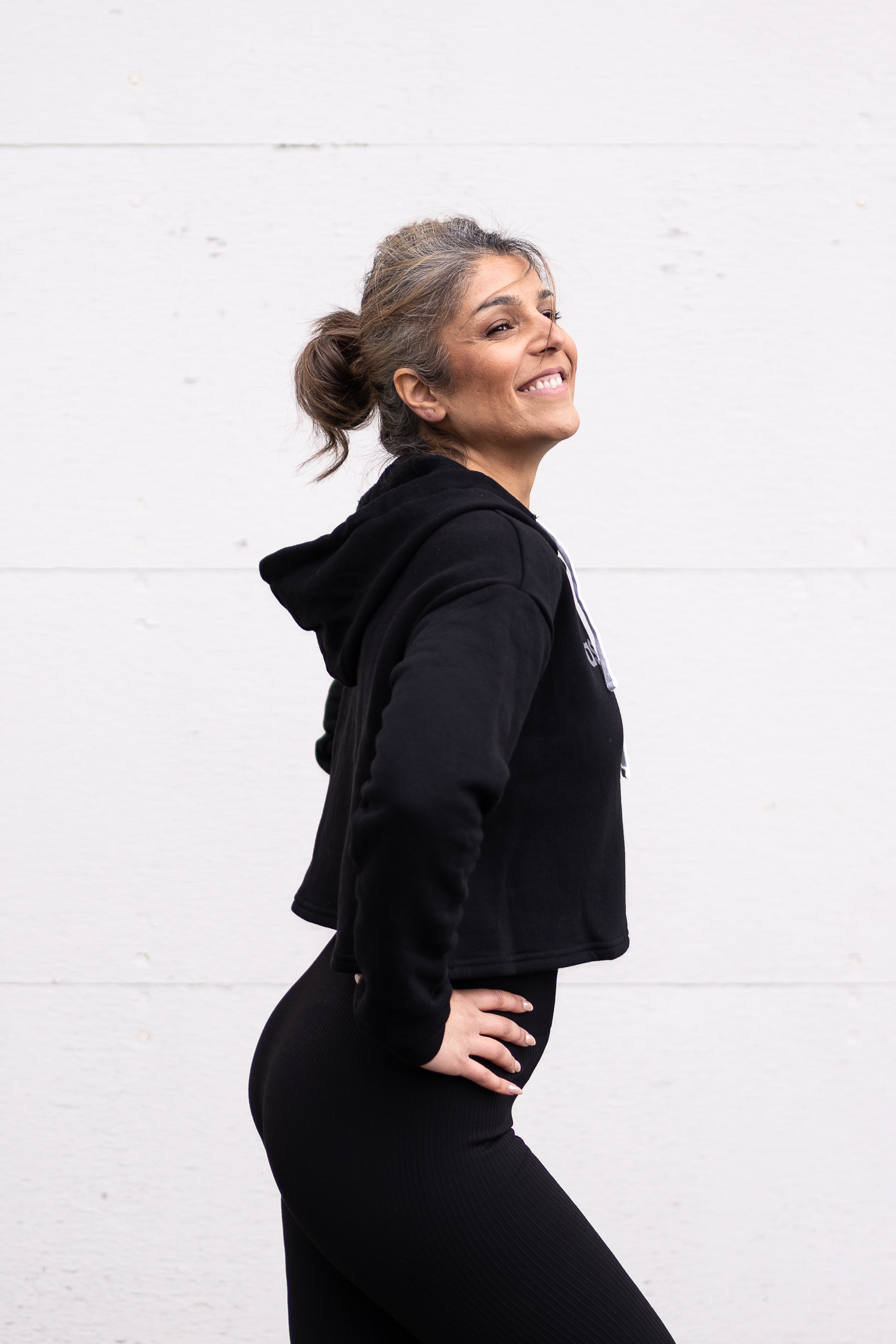 Woman standing facing sideways wearing a cropped bamboo-organic cotton fleece hoodie in black, featuring a finished hem, white drawstrings at the hood and the A$$piration Project logo on the front in light grey.