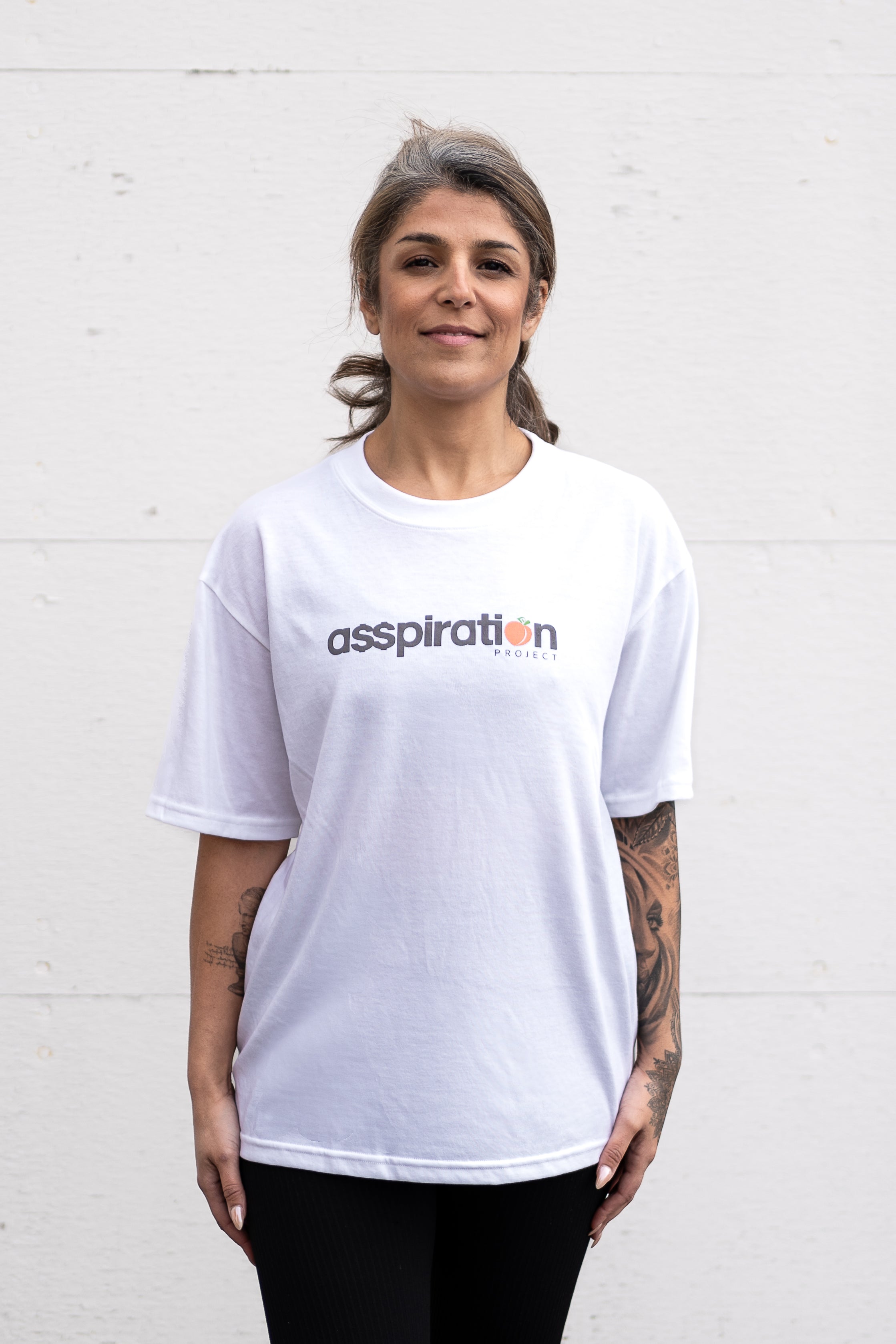 Woman standing facing forwards with arms at her sides, wearing a white unisex oversized t-shirt with a boxy fit made from premium poly-cotton jersey, featuring the A$$piration Project logo on the front in grey, peach and green.
