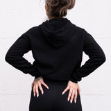 Woman standing facing sideways wearing a cropped bamboo-organic cotton fleece hoodie in black, featuring a finished hem.