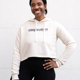Woman standing facing forward with her hand on her hip, wearing a cropped bamboo-organic cotton fleece hoodie in cream, featuring a finished hem, white drawstrings at the hood and the A$$piration Project logo on the front in grey, peach and green.