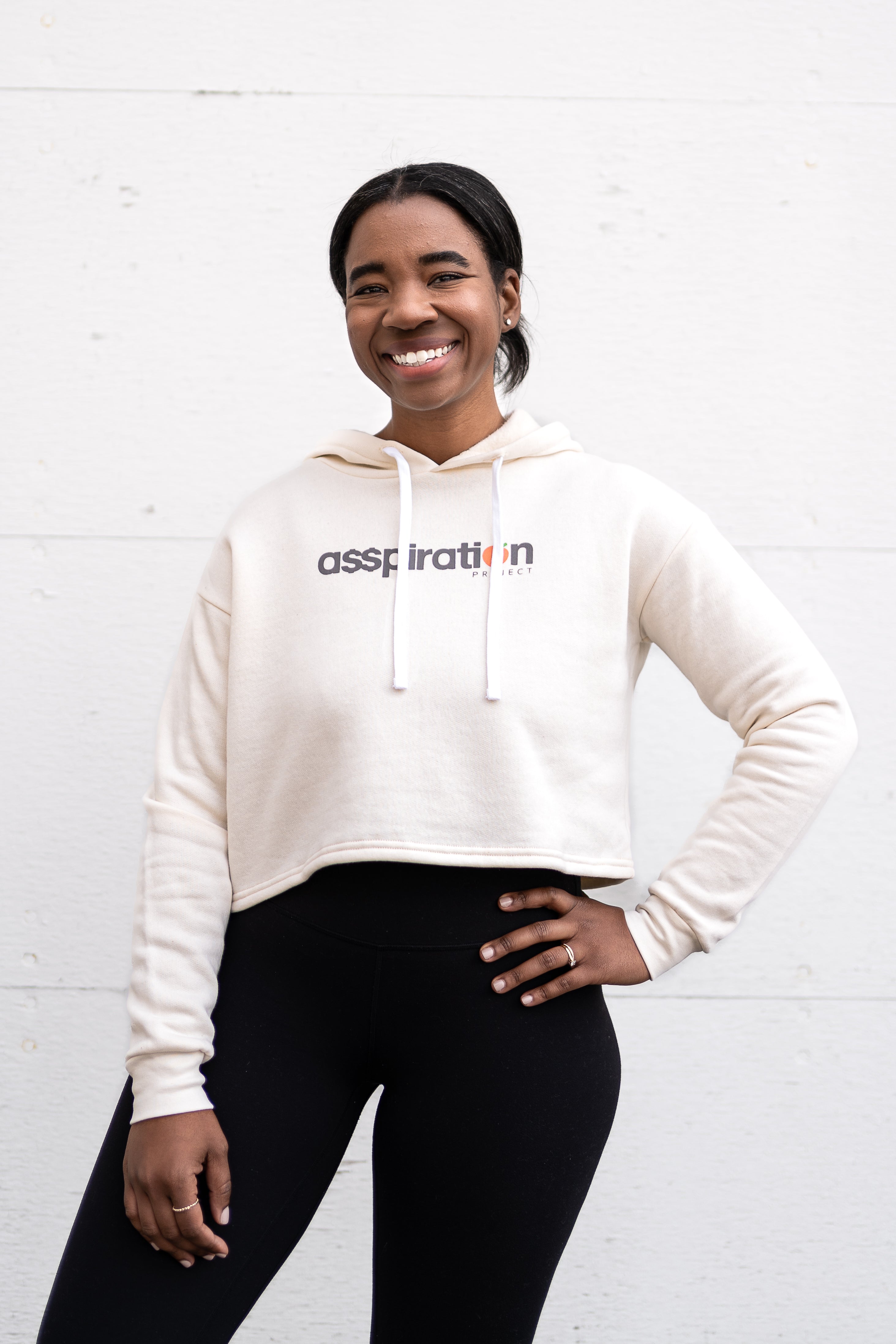 Woman standing facing forward with her hand on her hip, wearing a cropped bamboo-organic cotton fleece hoodie in cream, featuring a finished hem, white drawstrings at the hood and the A$$piration Project logo on the front in grey, peach and green.