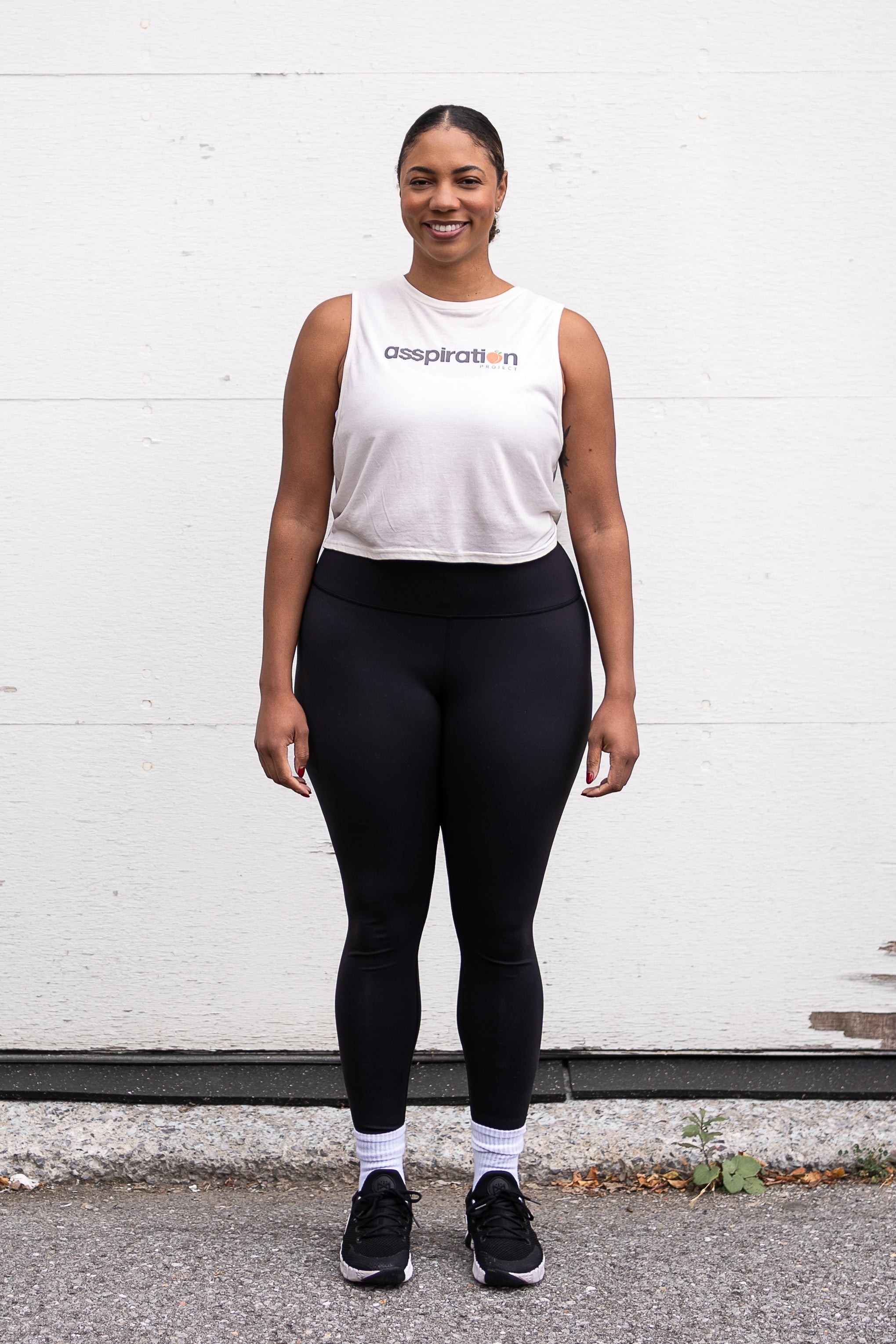 Woman standing facing forward with her arms by her sides, wearing a cropped bamboo-organic cotton tank in cream, featuring the A$$piration Project logo on the front in grey, peach and green.