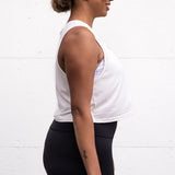 Woman standing facing sideways with her arms by her sides, wearing a cropped bamboo-organic cotton tank in cream, featuring the A$$piration Project logo on the front in grey, peach and green.