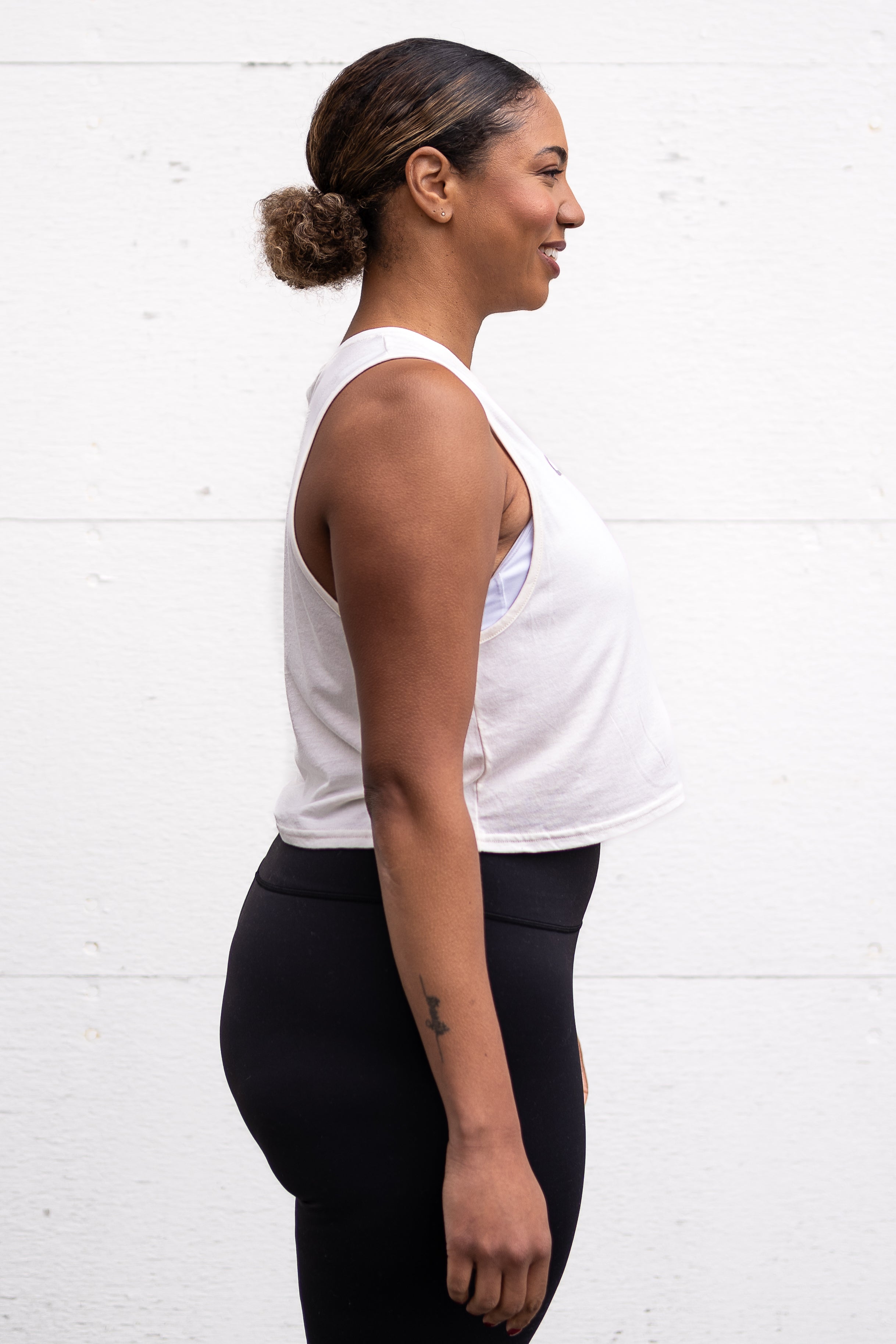 Woman standing facing sideways with her arms by her sides, wearing a cropped bamboo-organic cotton tank in cream, featuring the A$$piration Project logo on the front in grey, peach and green.