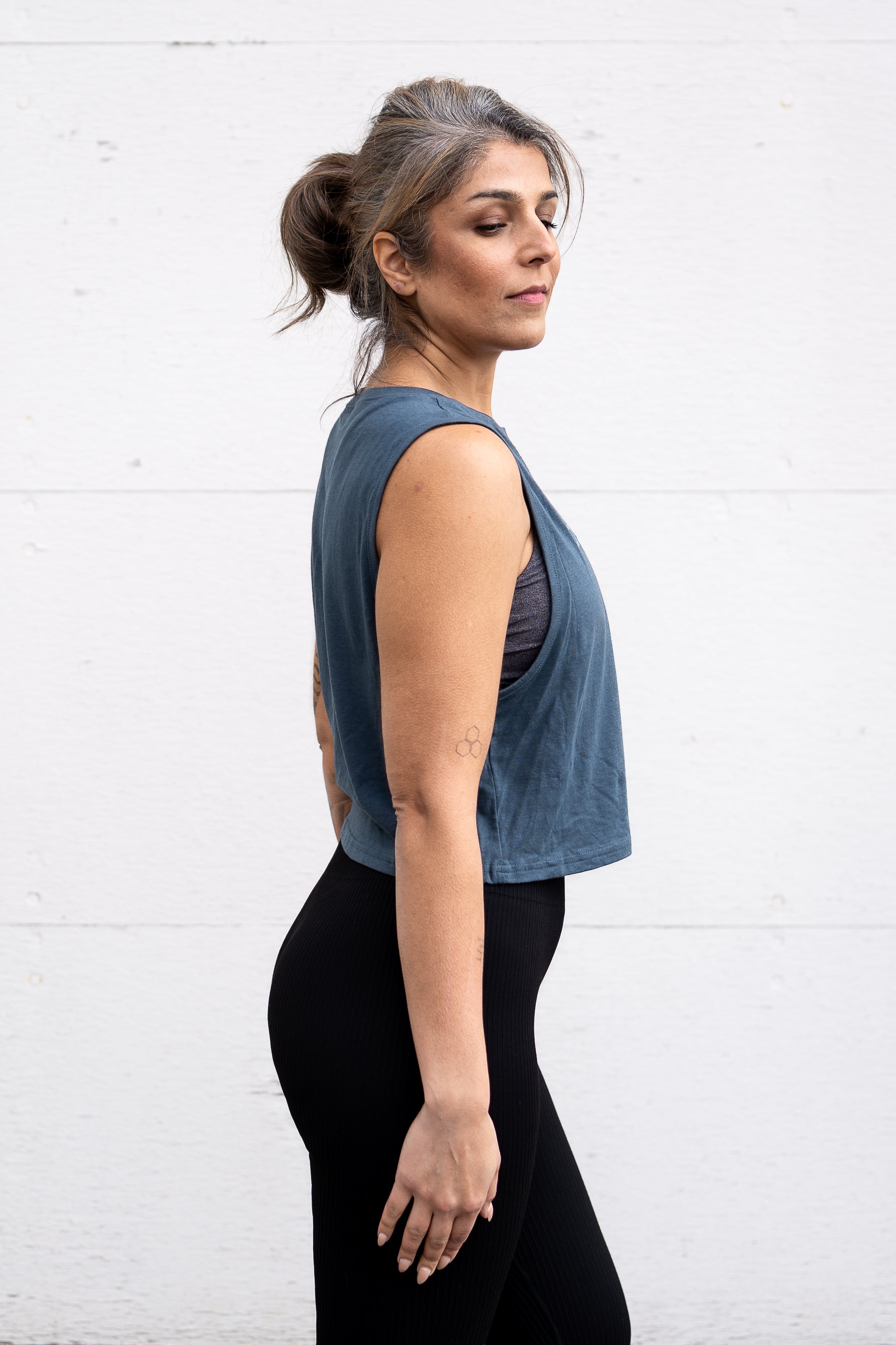 Woman standing facing sideways, wearing a cropped bamboo-organic cotton tank in slate blue, featuring the A$$piration Project logo on the front in light blue.