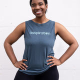 Woman standing facing forward with hands on her hips, wearing a full-length bamboo-organic cotton tank in slate blue, with a relaxed fit and rounded hem, featuring the A$$piration Project logo on the front in light blue.