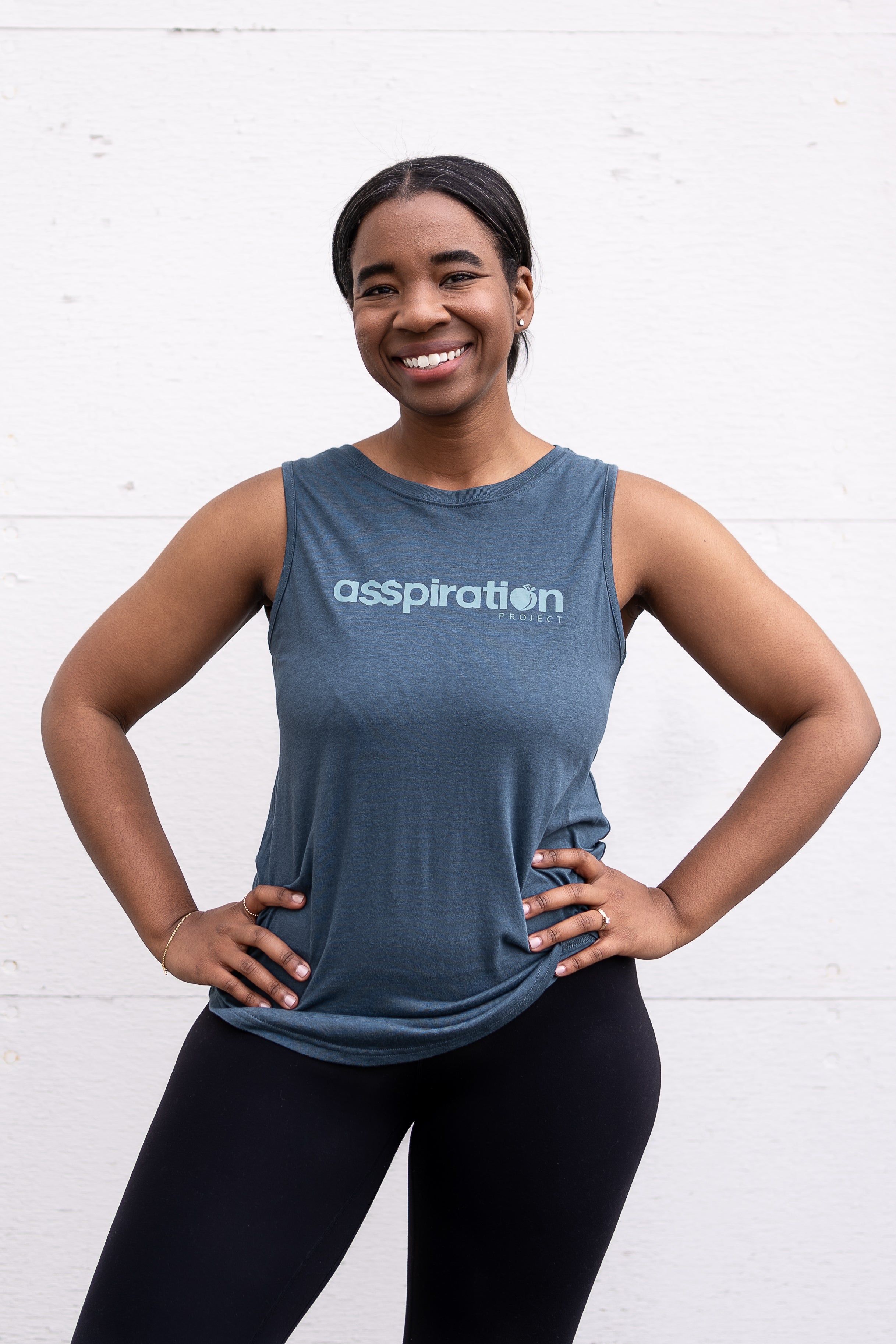Woman standing facing forward with hands on her hips, wearing a full-length bamboo-organic cotton tank in slate blue, with a relaxed fit and rounded hem, featuring the A$$piration Project logo on the front in light blue.