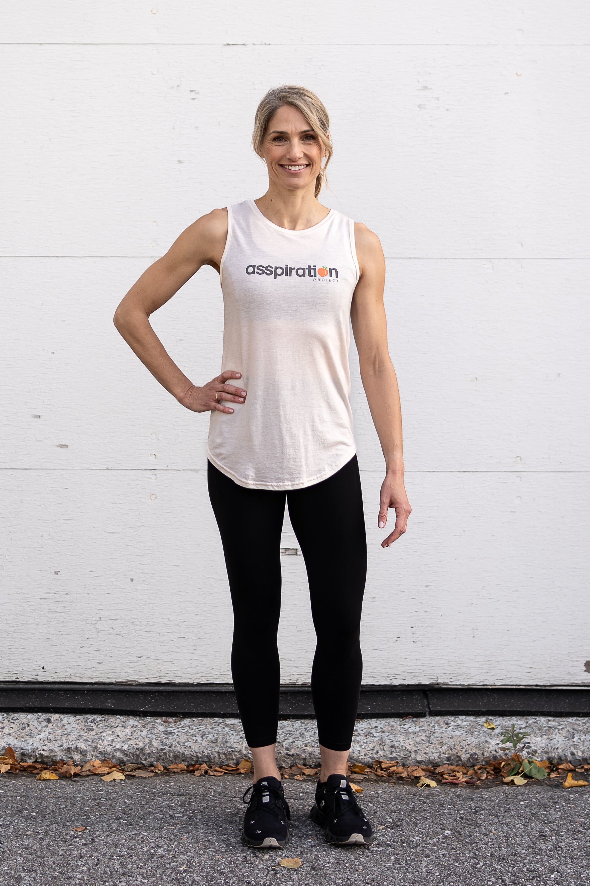 Woman standing facing forward with one hand on hip, wearing a full-length bamboo-organic cotton tank in cream, with a relaxed fit and rounded hem, featuring the A$$piration Project logo on the front in grey, peach and green.