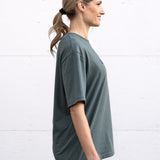 Woman standing facing sideways wearing a sage green unisex oversized t-shirt with a boxy fit, made from premium poly-cotton jersey, featuring the A$$piration Project logo on the front in a tone on tone darker sage green.