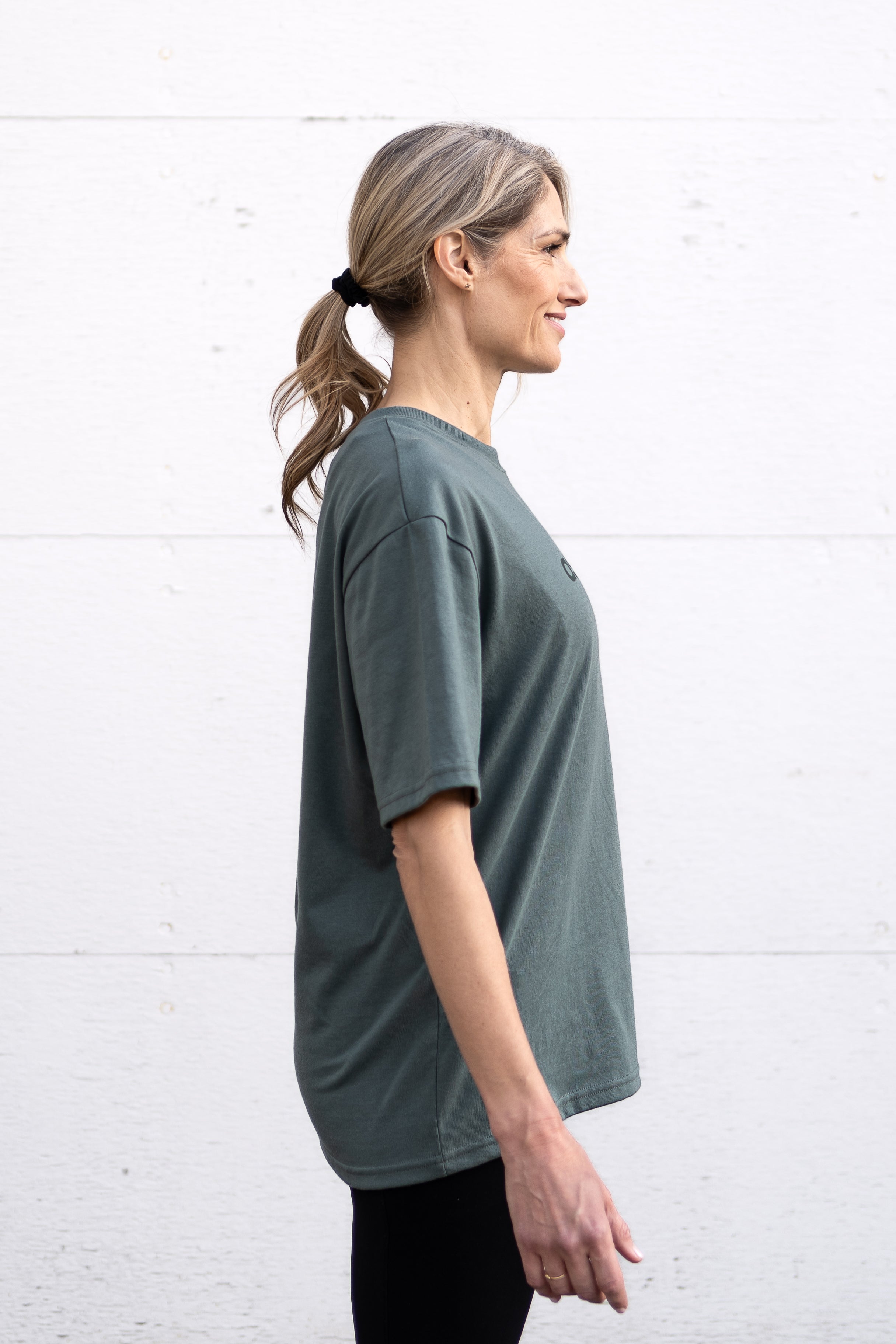 Woman standing facing sideways wearing a sage green unisex oversized t-shirt with a boxy fit, made from premium poly-cotton jersey, featuring the A$$piration Project logo on the front in a tone on tone darker sage green.
