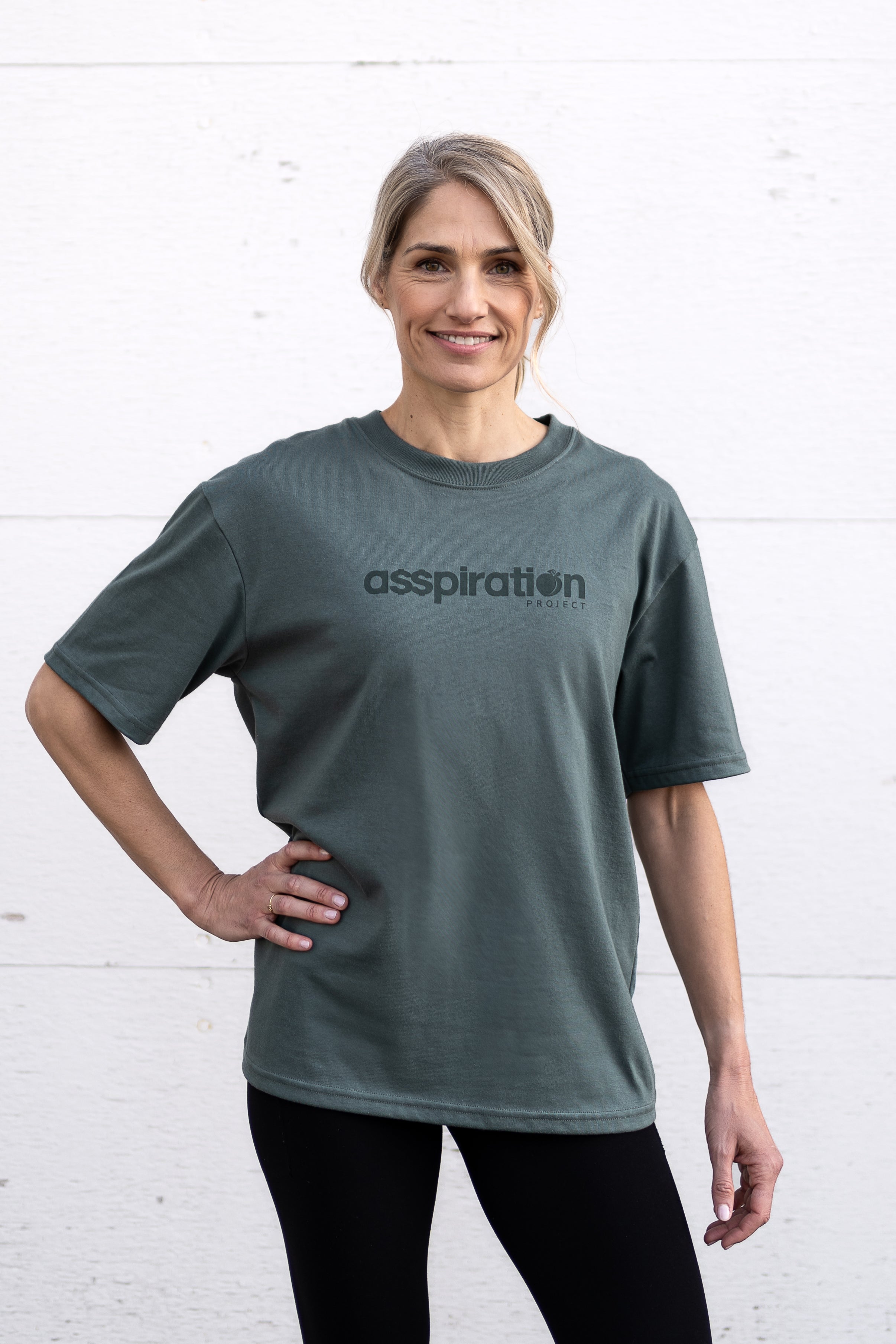 Woman standing facing forward with one hand on hip, wearing a sage green unisex oversized t-shirt with a boxy fit, made from premium poly-cotton jersey, featuring the A$$piration Project logo on the front in a tone on tone darker sage green.