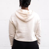 Woman standing facing backwards wearing a cropped bamboo-organic cotton fleece hoodie in cream, featuring a finished hem.