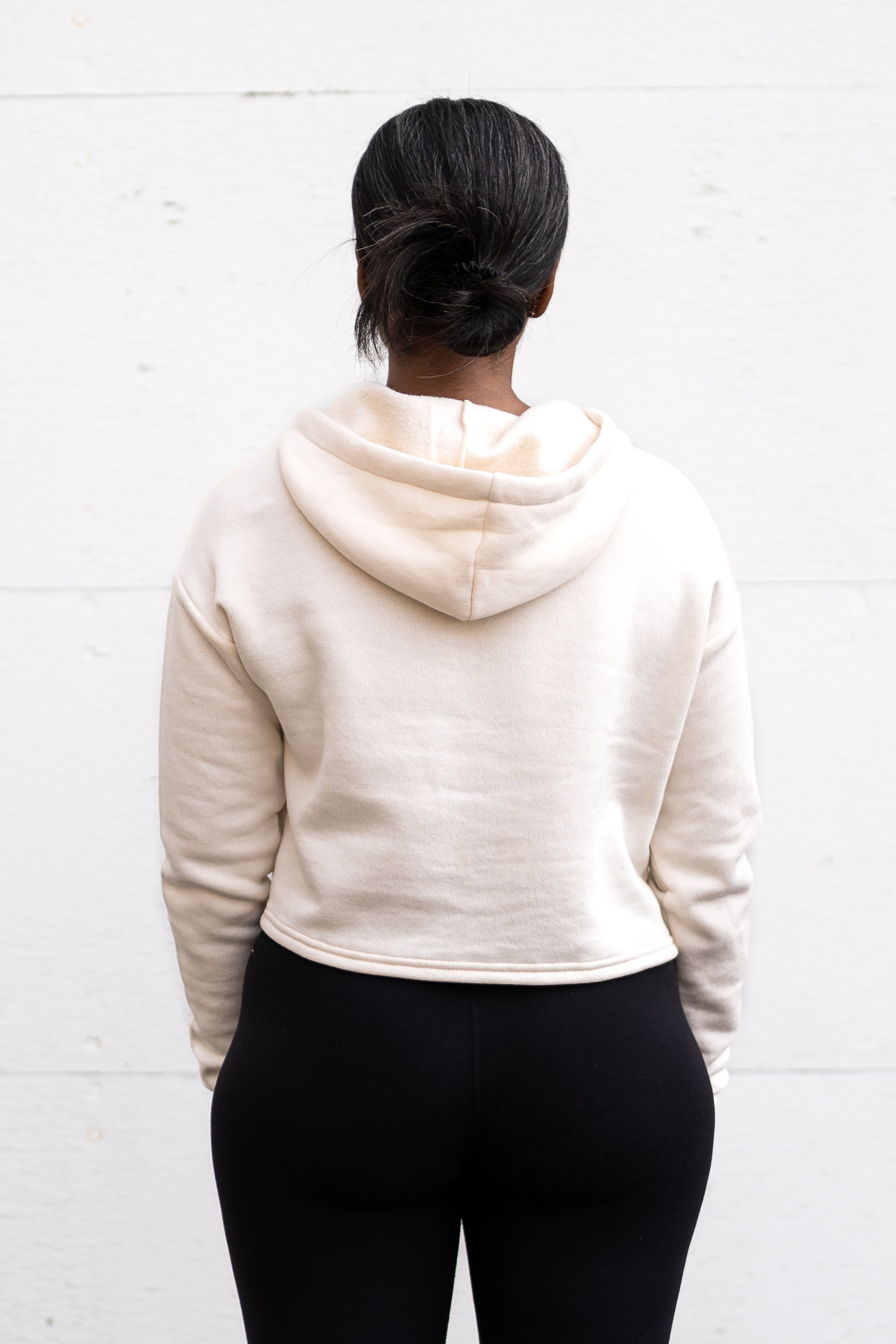 Woman standing facing backwards wearing a cropped bamboo-organic cotton fleece hoodie in cream, featuring a finished hem.
