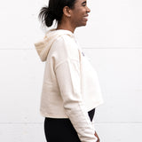 Woman standing facing sideways wearing a cropped bamboo-organic cotton fleece hoodie in cream, featuring a finished hem, white drawstrings at the hood and the A$$piration Project logo on the front in grey, peach and green.