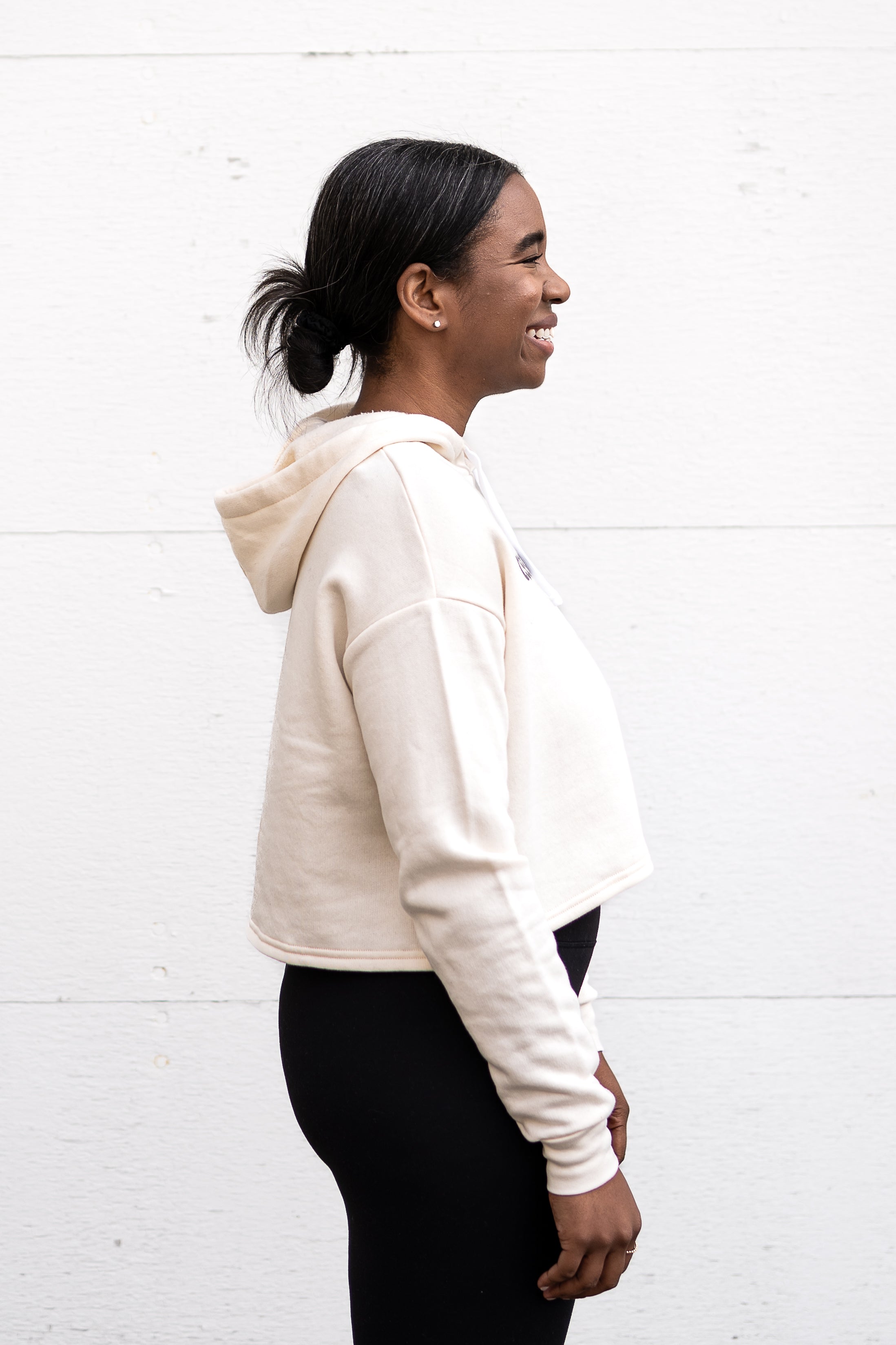 Woman standing facing sideways wearing a cropped bamboo-organic cotton fleece hoodie in cream, featuring a finished hem, white drawstrings at the hood and the A$$piration Project logo on the front in grey, peach and green.
