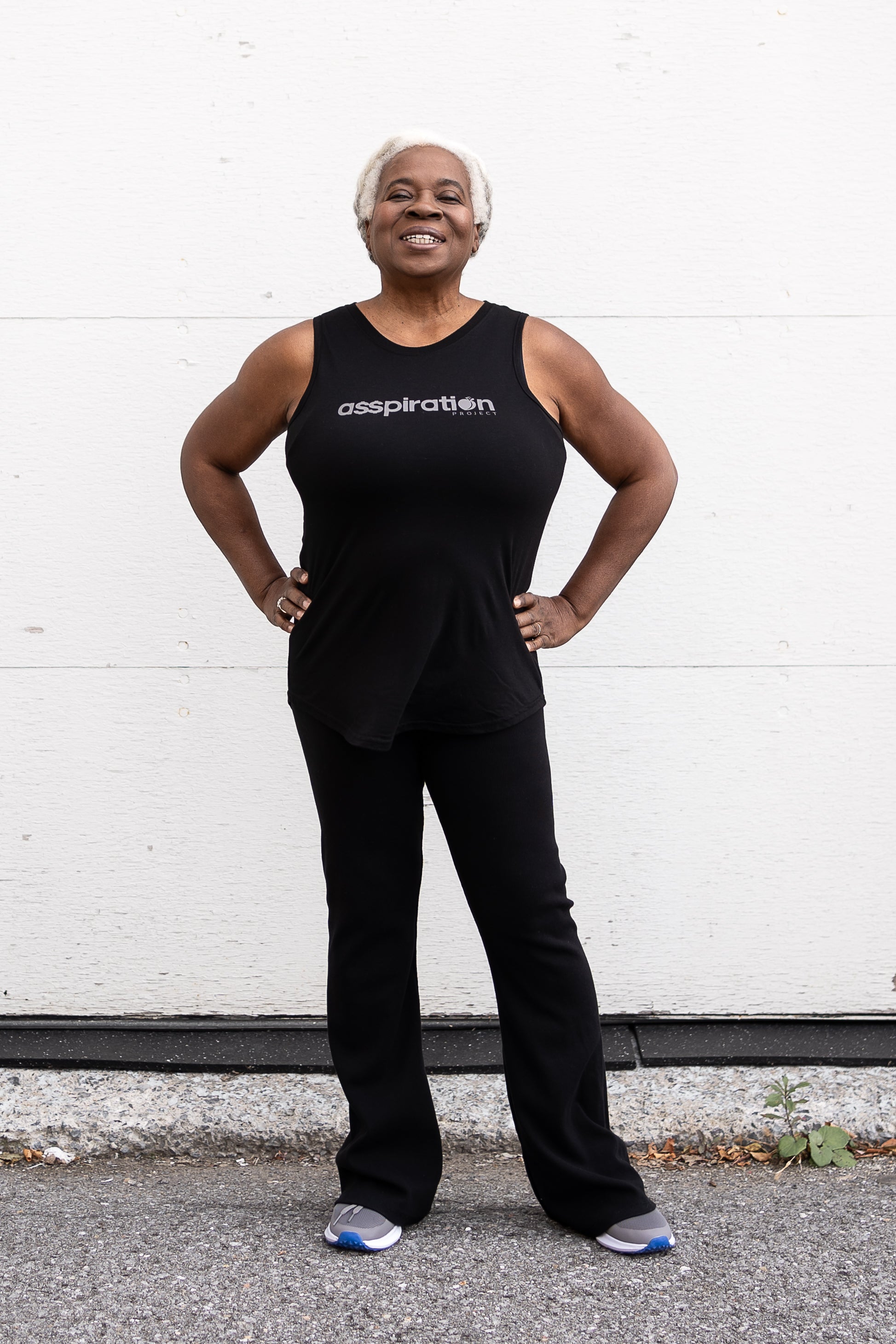 Woman standing facing forward with her hands on her hips, wearing a full-length bamboo-organic cotton tank in black, with a relaxed fit and rounded hem, featuring the A$$piration Project logo on the front in light grey.