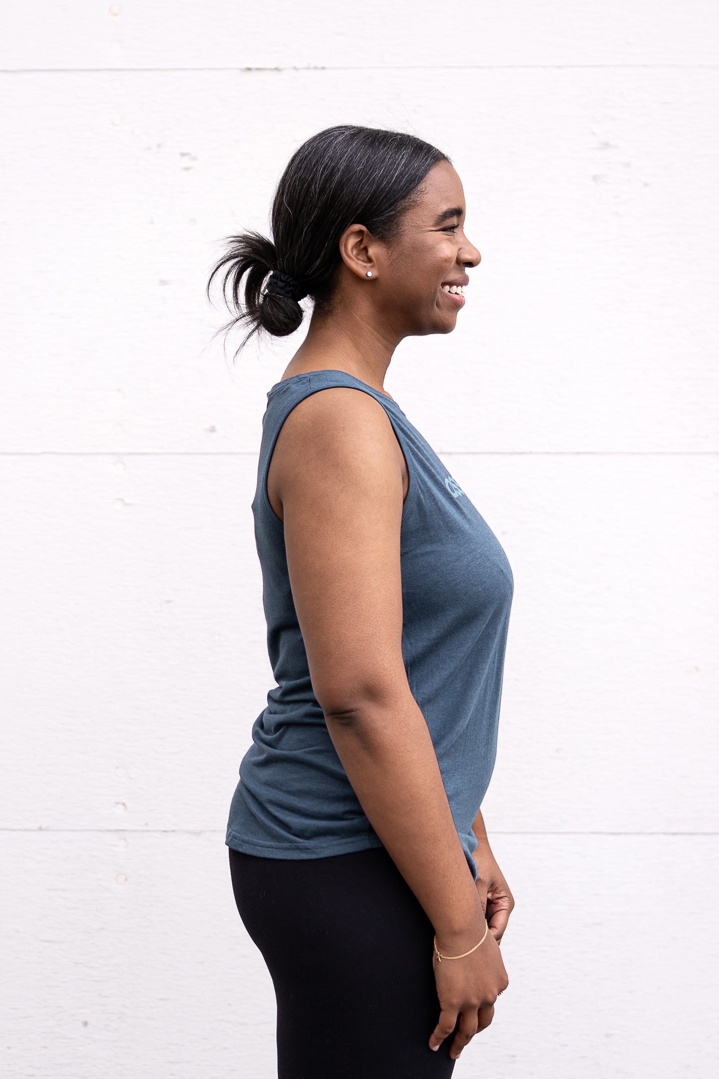 Woman standing in side profile wearing a full-length bamboo-organic cotton tank in slate blue, with a relaxed fit and rounded hem, featuring the A$$piration Project logo on the front in light blue.