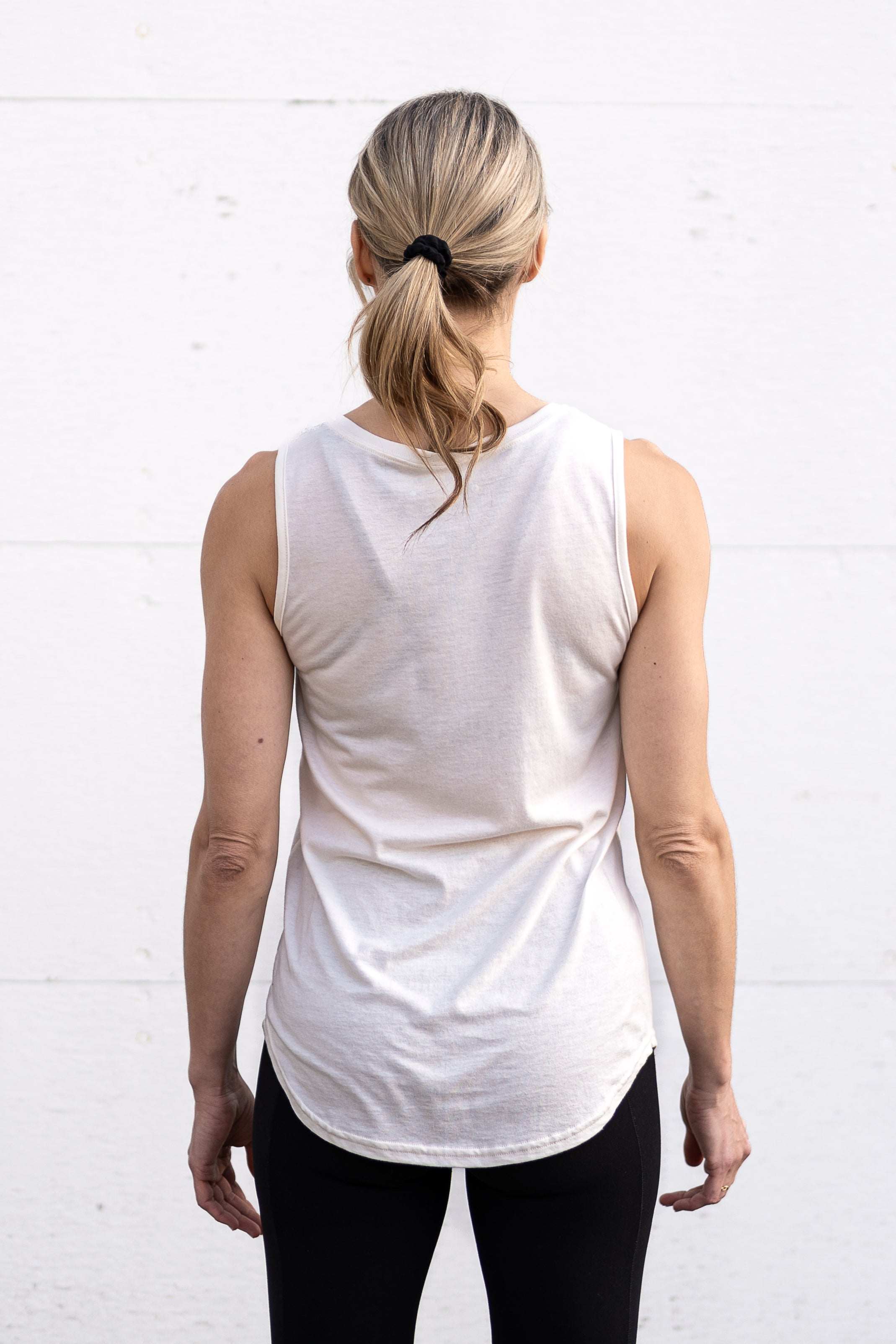 Woman standing facing backwards wearing a full-length bamboo-organic cotton tank in cream, featuring a relaxed fit and rounded hem.