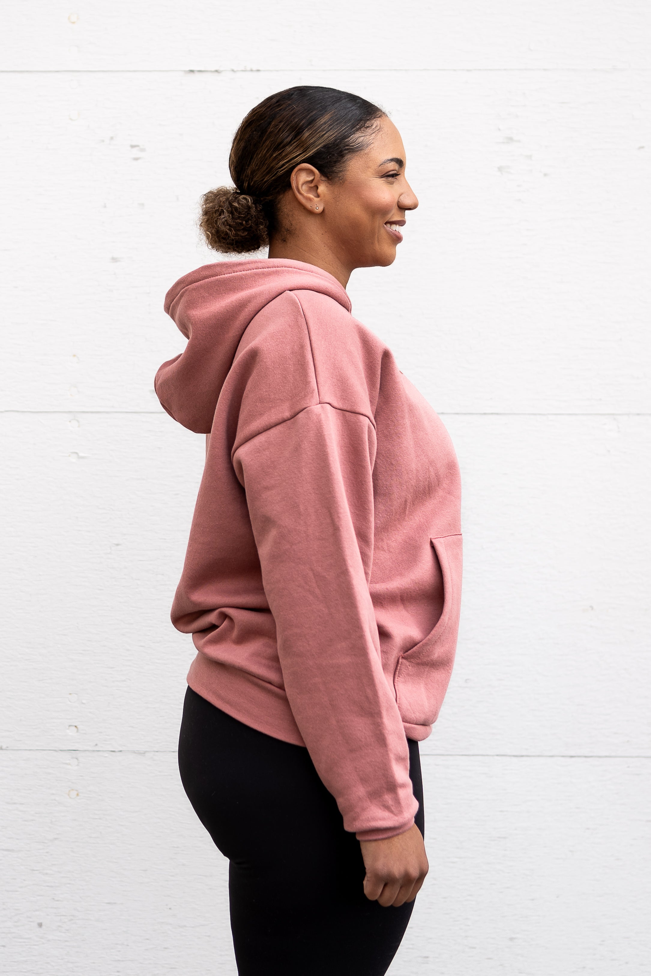 Woman standing facing sideways wearing a unisex oversize bamboo-organic cotton fleece hoodie in dusty rose, with dropped shoulders and a large kangaroo pocket, featuring the A$$piration Project logo on the front in tone on tone dark ruby pink.