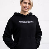 Woman standing facing forward with hands inside her front pocket, wearing a unisex oversize bamboo-organic cotton fleece hoodie in black, with dropped shoulders and a large kangaroo pocket, featuring the A$$piration Project logo on the front in light grey.