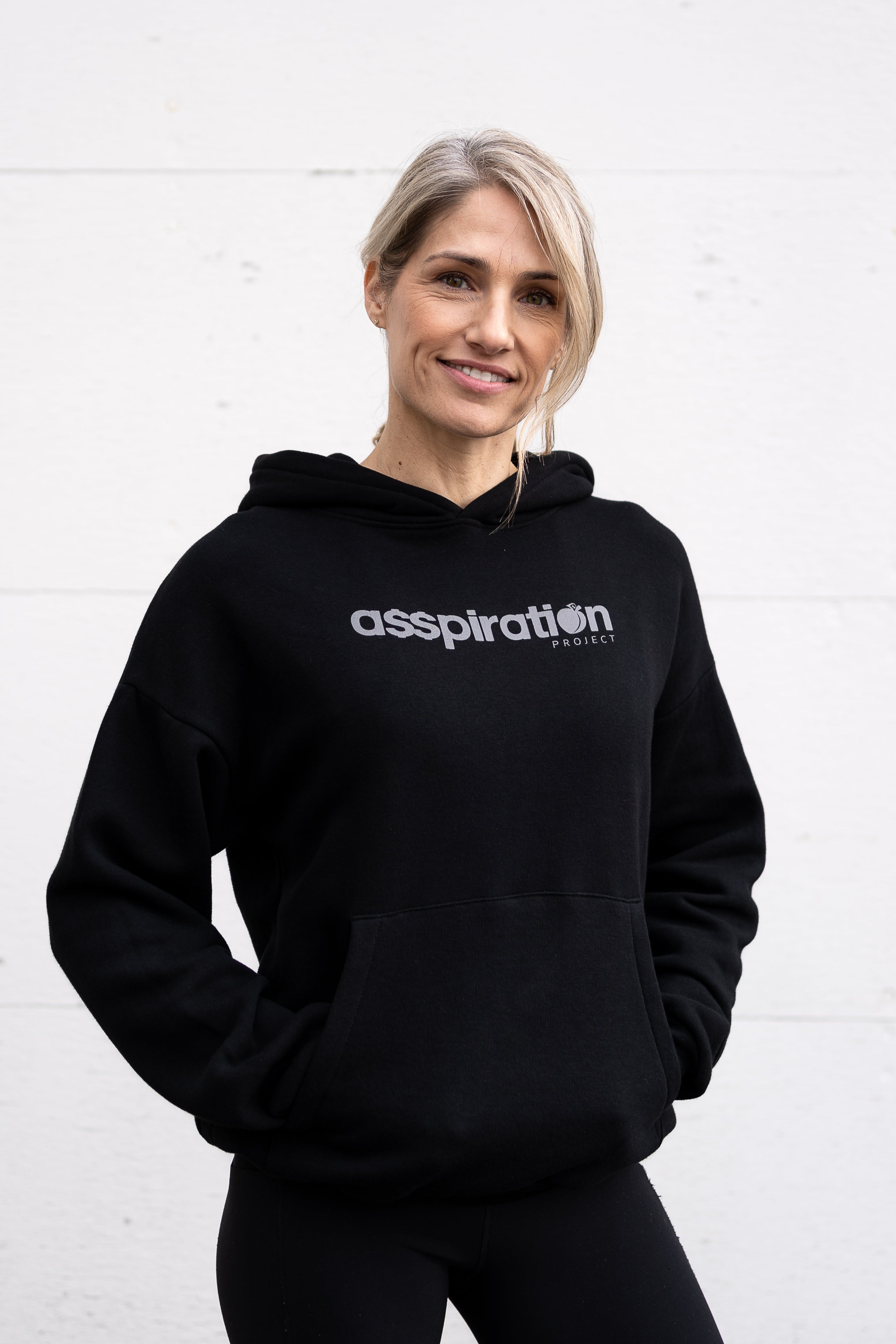 Woman standing facing forward with hands inside her front pocket, wearing a unisex oversize bamboo-organic cotton fleece hoodie in black, with dropped shoulders and a large kangaroo pocket, featuring the A$$piration Project logo on the front in light grey.