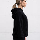 Woman standing facing sideways wearing a unisex oversize bamboo-organic cotton fleece hoodie in black, with dropped shoulders and a large kangaroo pocket, featuring the A$$piration Project logo on the front in light grey.