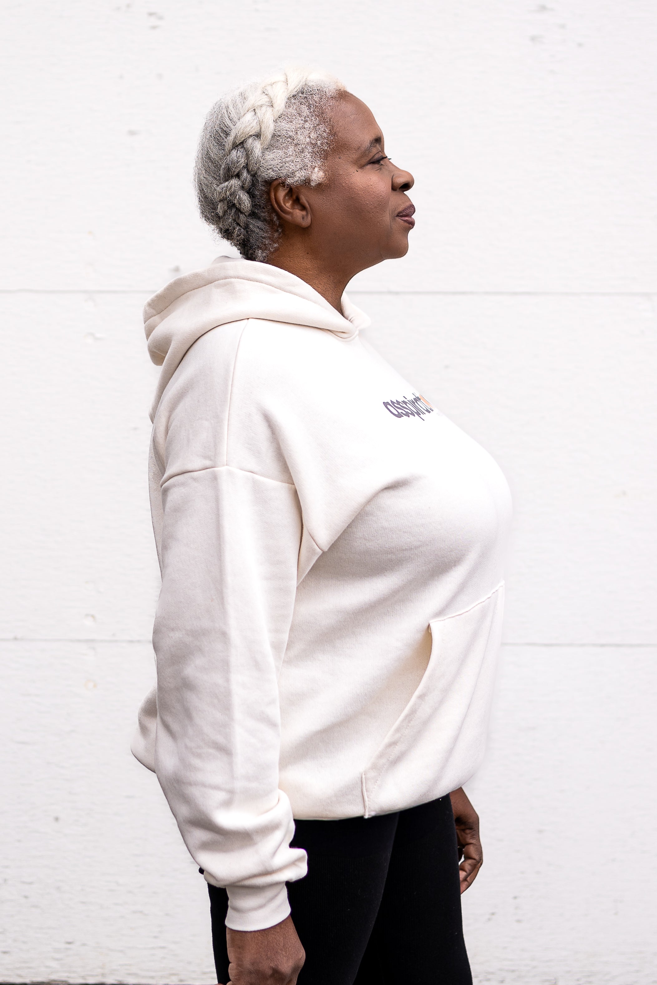 Woman standing facing sideways with arms at her sides, wearing a unisex oversize bamboo-organic cotton fleece hoodie in cream, with dropped shoulders and a large kangaroo pocket, featuring the A$$piration Project logo on the front in grey, peach and green.