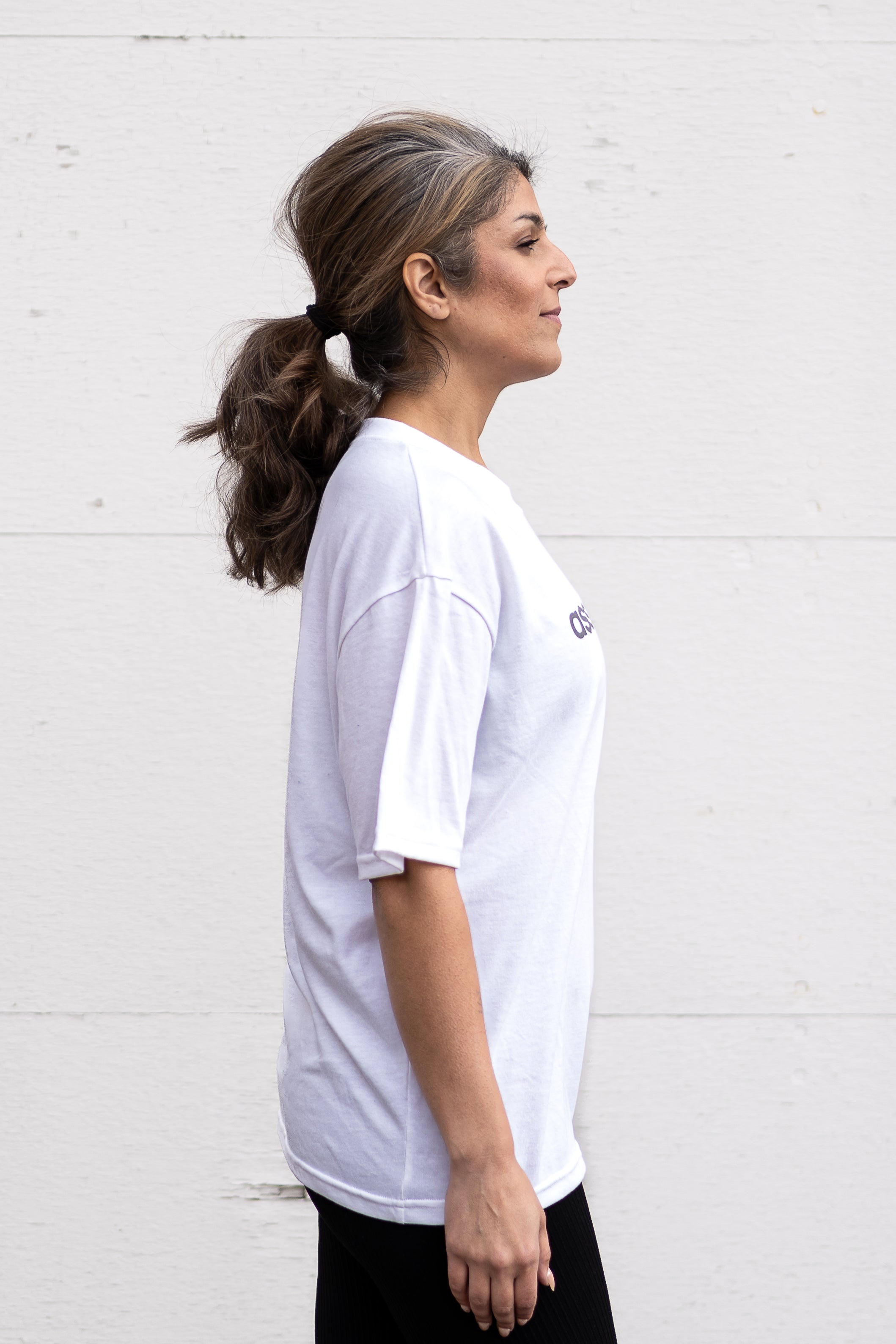 Woman standing facing sideways wearing a white unisex oversized t-shirt with a boxy fit made from premium poly-cotton jersey, featuring the A$$piration Project logo on the front in grey, peach and green.
