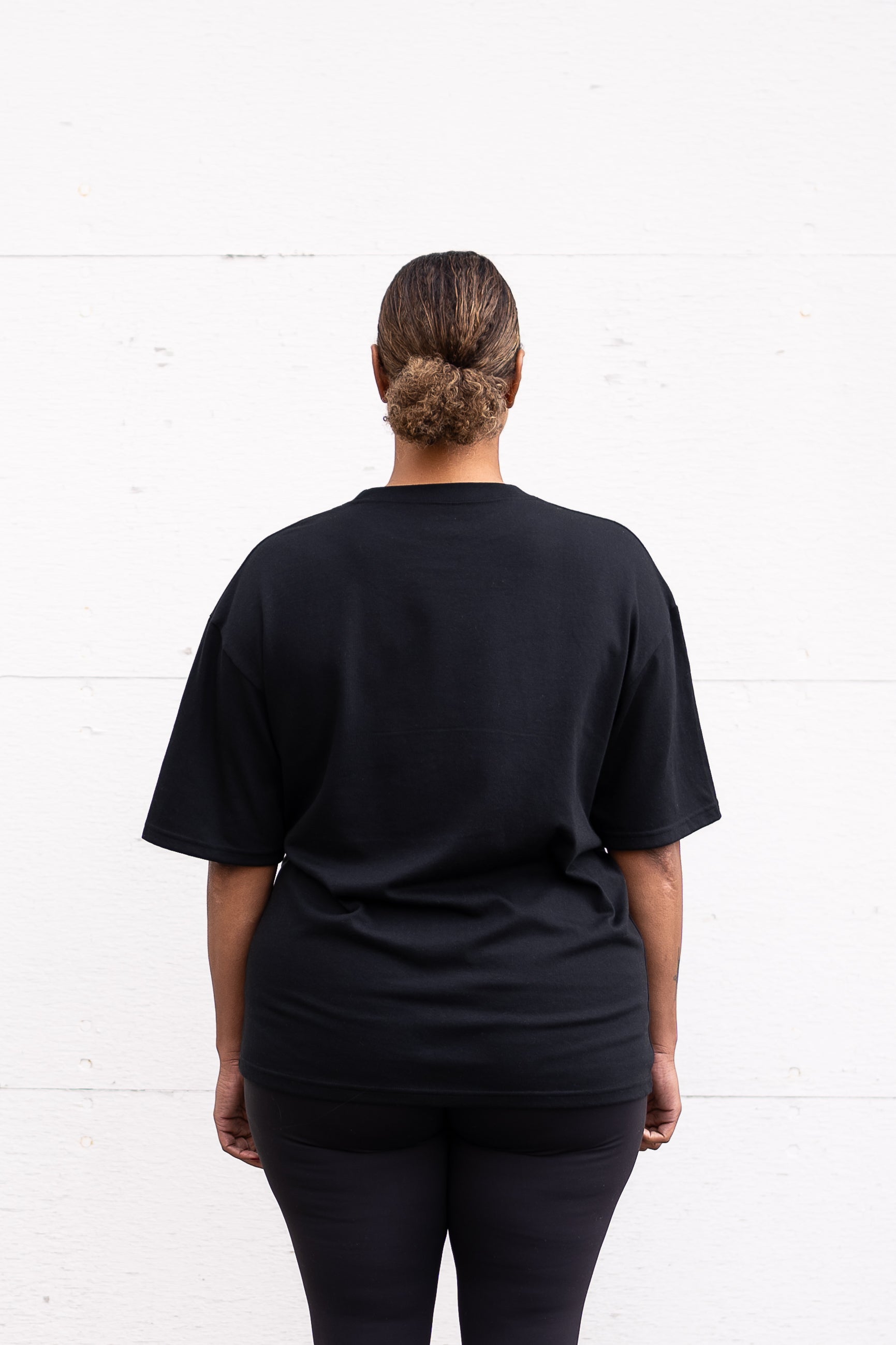 Woman standing facing backwards wearing a black unisex oversized t-shirt with a boxy fit, made from premium poly-cotton jersey.