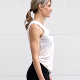 Woman facing sideways wearing a full-length bamboo-organic cotton tank in cream, displaying the back hem tucked into her sports bra, and the front hem rounded at the waist, featuring the A$$piration Project logo on the front in grey, peach and green.