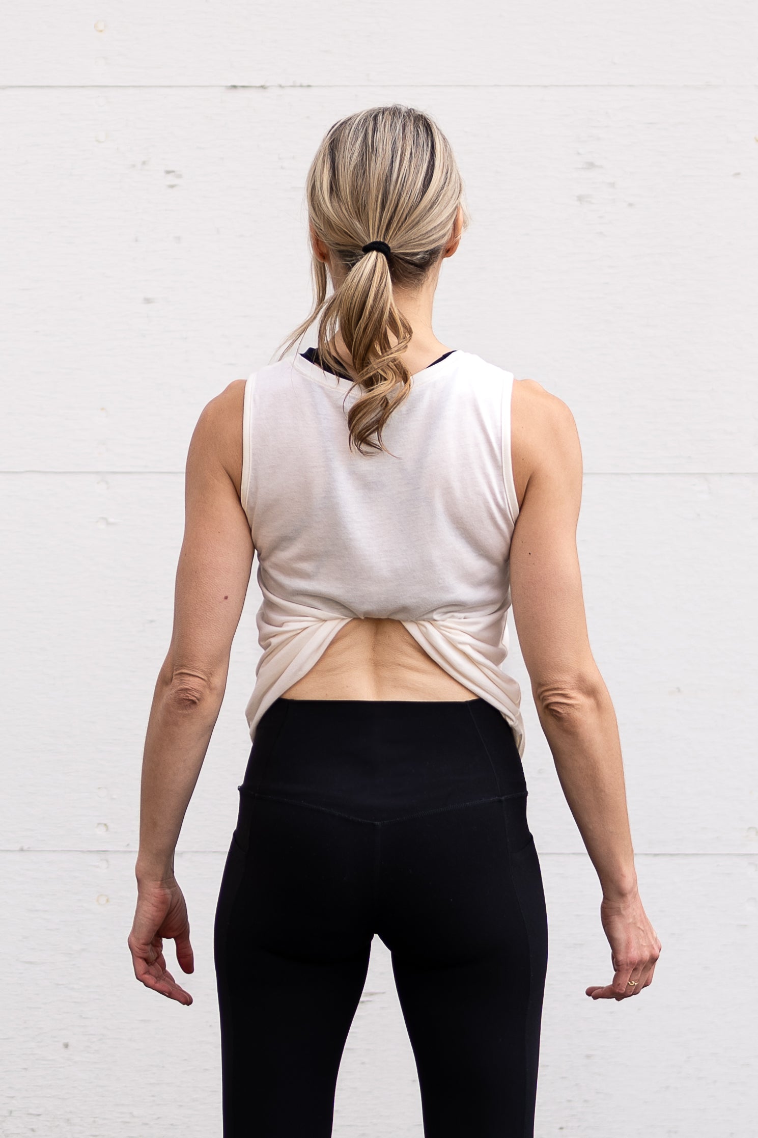 Woman standing facing backwards wearing a full-length bamboo-organic cotton tank in cream, displaying the back hem tucked into her sports bra.