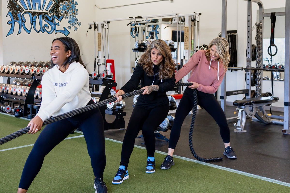Three women in a gym engaged in "tug of war" with battle ropes, wearing cropped bamboo-organic cotton hoodies in cream, black and dusty rose, all featuring the A$$piration Project logo on the front.