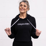 Woman standing facing forward wearing a cropped bamboo-organic cotton fleece hoodie in black, featuring a finished hem, white drawstrings at the hood and the A$$piration Project logo on the front in light grey.