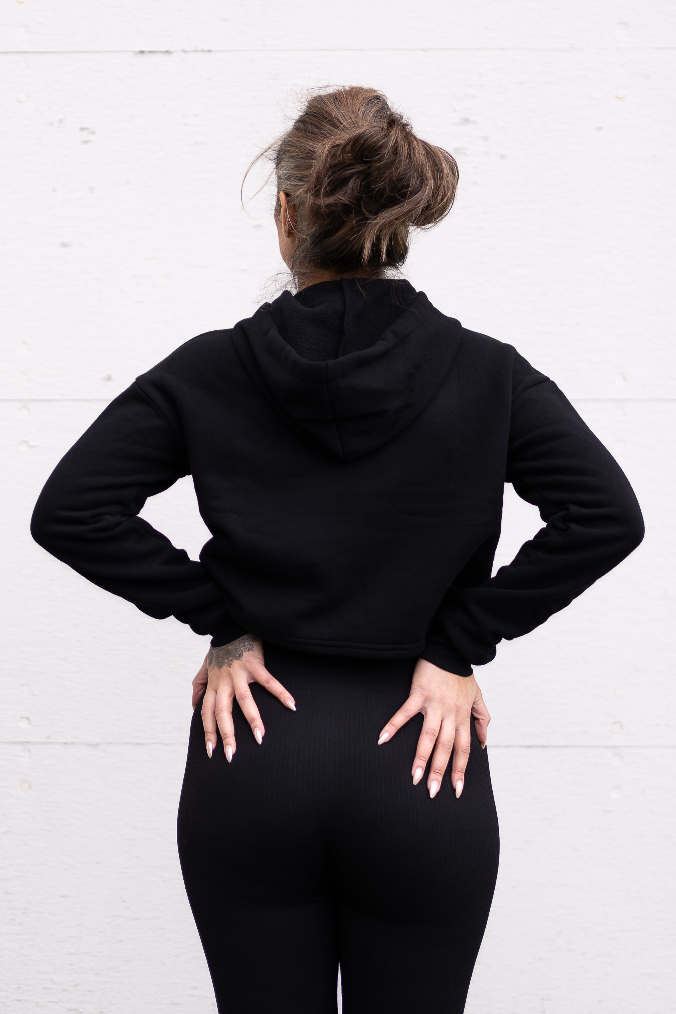 Woman standing facing sideways wearing a cropped bamboo-organic cotton fleece hoodie in black, featuring a finished hem.