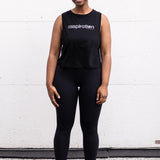 Woman standing facing forward with her arms by her sides, wearing a cropped bamboo-organic cotton tank in black, featuring the A$$piration Project logo on the front in light grey.