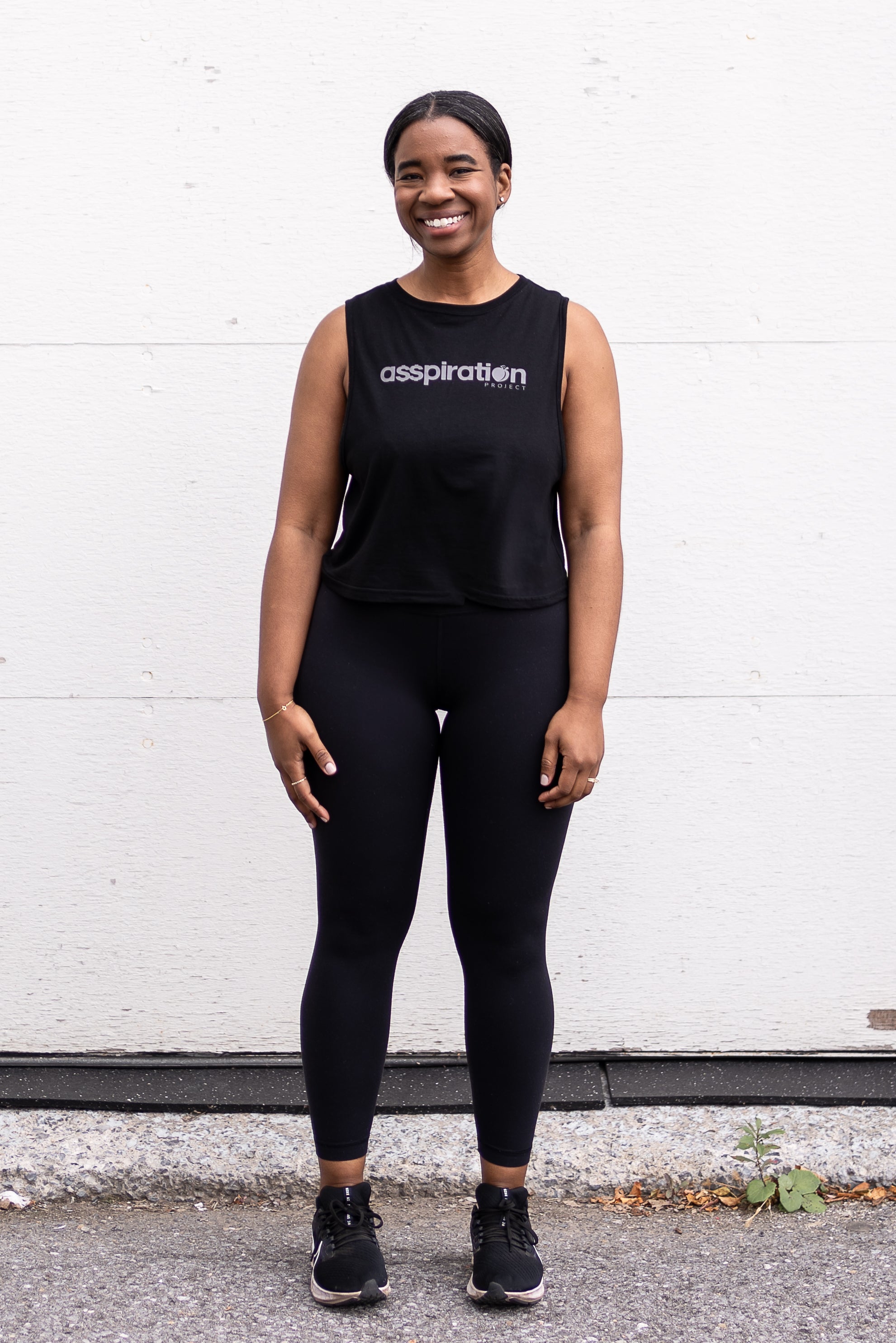 Woman standing facing forward with her arms by her sides, wearing a cropped bamboo-organic cotton tank in black, featuring the A$$piration Project logo on the front in light grey.