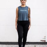 Woman standing facing forward with her arms by her sides, wearing a cropped bamboo-organic cotton tank in slate blue, featuring the A$$piration Project logo on the front in light blue.