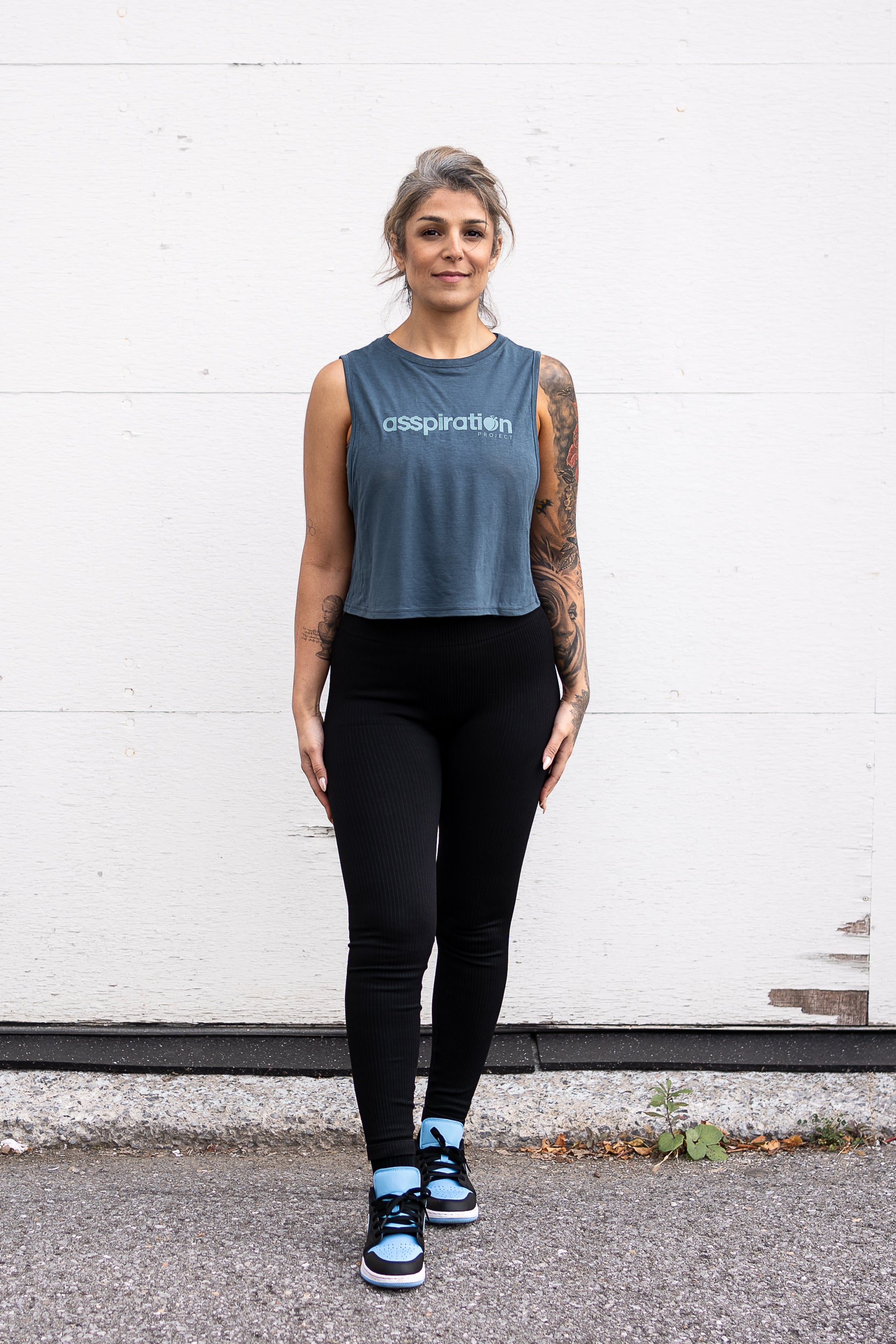 Woman standing facing forward with her arms by her sides, wearing a cropped bamboo-organic cotton tank in slate blue, featuring the A$$piration Project logo on the front in light blue.