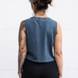 Woman standing facing backwards with her arms by her sides, wearing a cropped bamboo-organic cotton tank in slate blue.