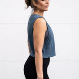 Woman standing facing sideways, wearing a cropped bamboo-organic cotton tank in slate blue, featuring the A$$piration Project logo on the front in light blue.