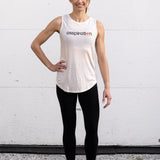 Woman standing facing forward with one hand on hip, wearing a full-length bamboo-organic cotton tank in cream, with a relaxed fit and rounded hem, featuring the A$$piration Project logo on the front in grey, peach and green.