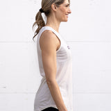 Woman wearing facing sideways in a full-length bamboo-organic cotton tank in cream, with a relaxed fit and rounded hem, featuring the A$$piration Project logo on the front in grey, peach and green.