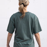 Woman standing facing backwards wearing a sage green unisex oversized t-shirt with a boxy fit, made from premium poly-cotton jersey.