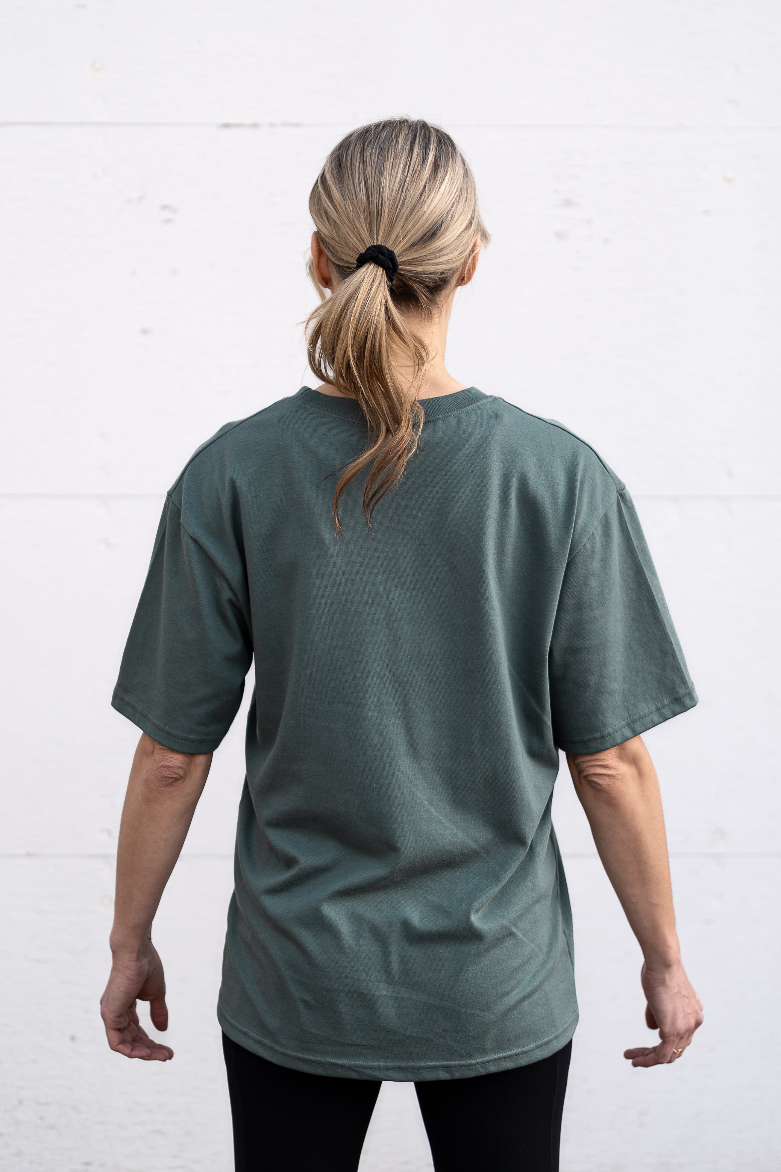 Woman standing facing backwards wearing a sage green unisex oversized t-shirt with a boxy fit, made from premium poly-cotton jersey.