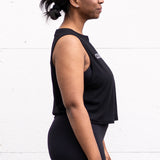 Woman standing facing sideways with her arms by her sides, wearing a cropped bamboo-organic cotton tank in black, featuring the A$$piration Project logo on the front in light grey.
