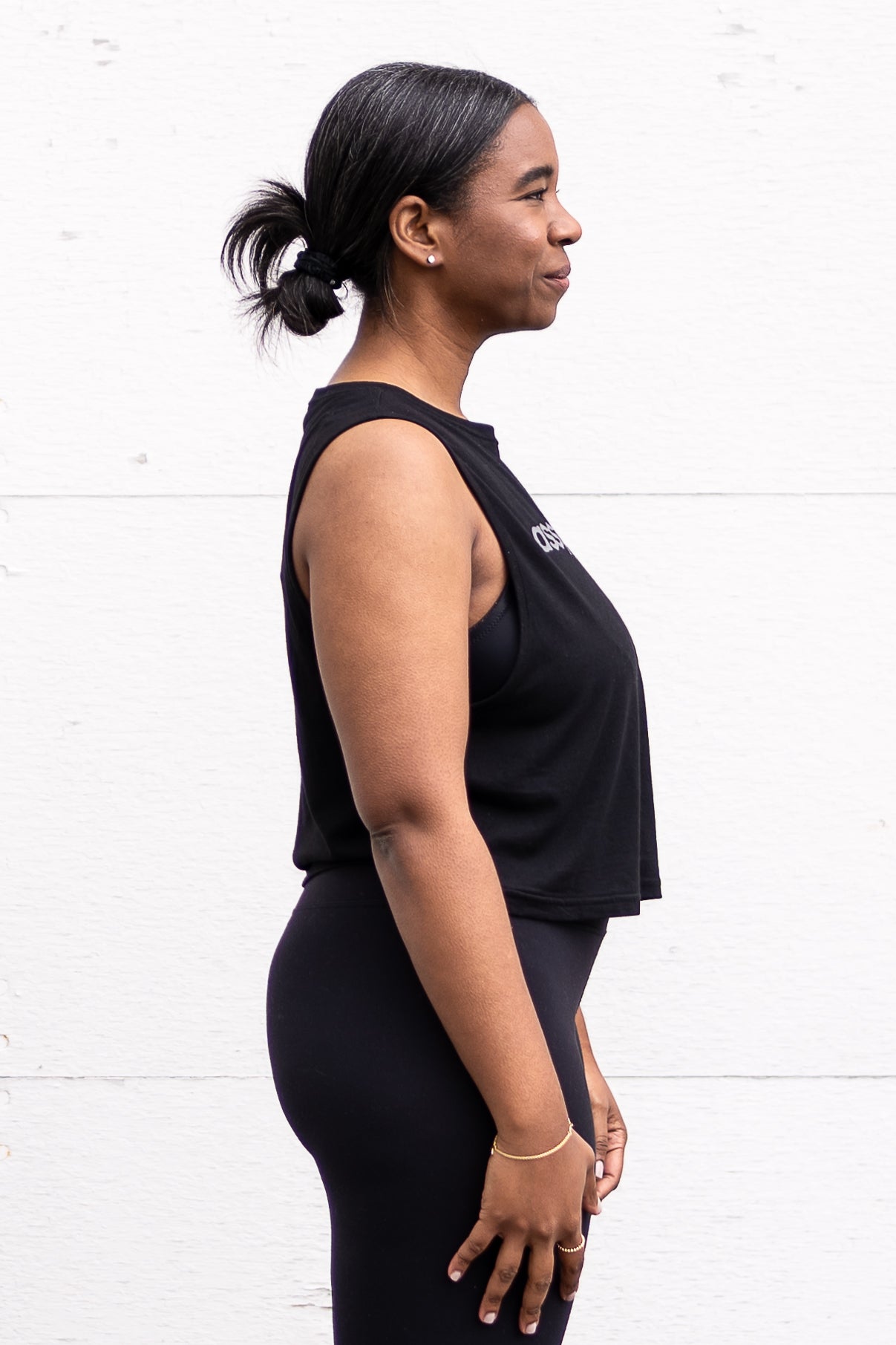 Woman standing facing sideways with her arms by her sides, wearing a cropped bamboo-organic cotton tank in black, featuring the A$$piration Project logo on the front in light grey.