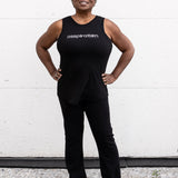 Woman standing facing forward with her hands on her hips, wearing a full-length bamboo-organic cotton tank in black, with a relaxed fit and rounded hem, featuring the A$$piration Project logo on the front in light grey.