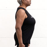 Woman standing facing sideways wearing a full-length bamboo-organic cotton tank in black, featuring a relaxed fit and rounded hem, featuring the A$$piration Project logo on the front in light grey.