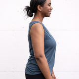 Woman standing in side profile wearing a full-length bamboo-organic cotton tank in slate blue, with a relaxed fit and rounded hem,  featuring the A$$piration Project logo on the front in light blue.